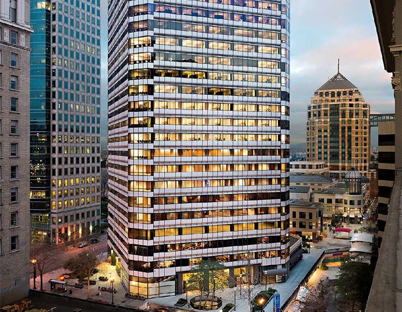 A lit-up modern office building at dusk, surrounded by other urban high-rise structures.