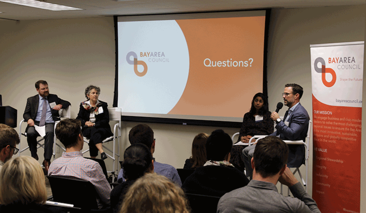 A panel discussion in progress with four speakers seated at the front of a room, addressing an audience.